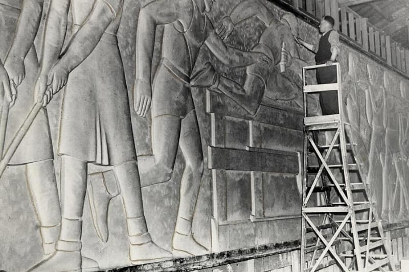 A black-and-white photo of a man standing on a tall wooden ladder and working on an elaborate wall sculpture.