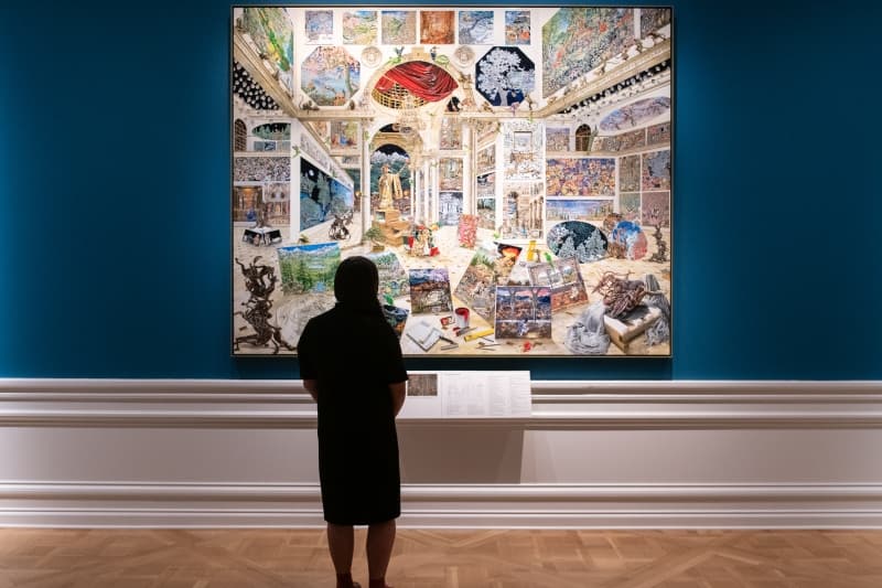 A person in shadow stands in front of a large, vibrant painting depicting an opulent and cavernous room filled with artworks.