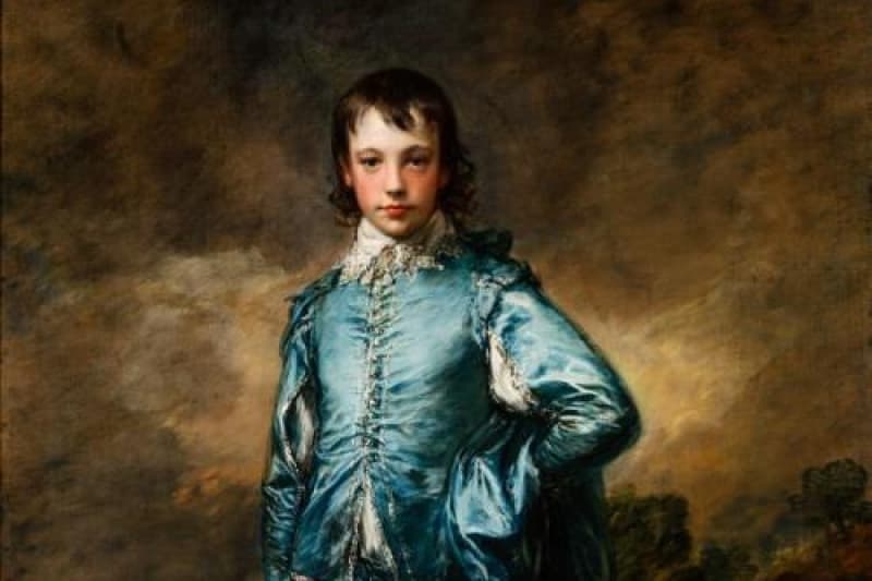 A full-length boy standing in a blue silky costume with a hand on his hip, his left leg forward, and a hat in his right hand set against a gray-brown background. 