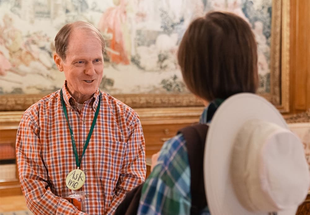 A person wearing an "Ask Me" badge talks to another person in front of a large hanging tapestry.