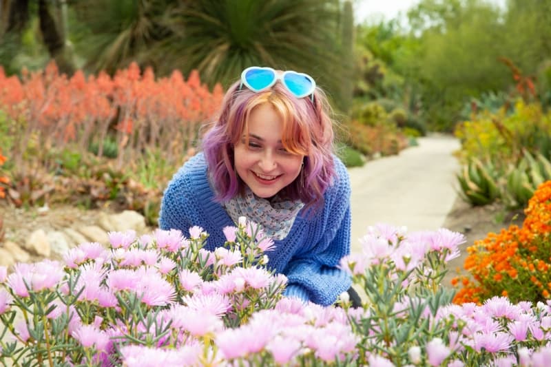 A smiling person with pink hair, blue glasses, and a blue sweater, kneels to look at pink flowers in a desert-themed garden.