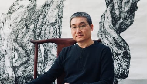 A person sits in a wood chair in front of a wall-sized painting.